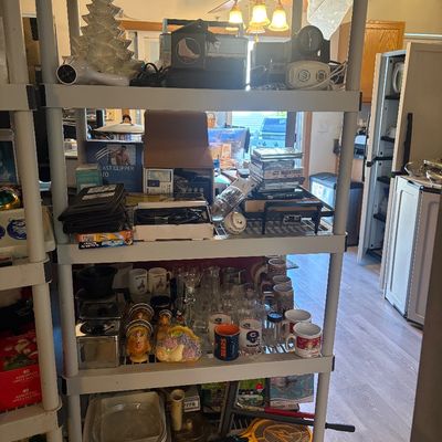 Sale Photo Thumbnail #57: Four-tier plastic shelving unit loaded with various household items, including glassware, ceramic mugs, small appliances, media cases, and decorative accents.