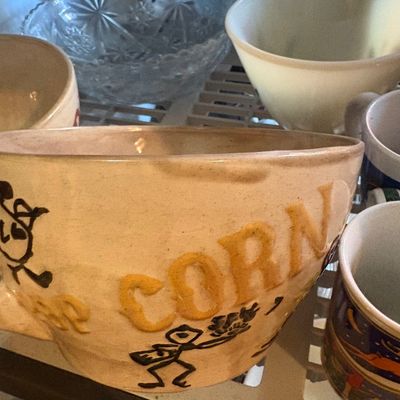 Sale Photo Thumbnail #84: Vintage ceramic popcorn bowl featuring raised lettering and stick figure illustrations, sitting among other kitchen glassware and dishware.