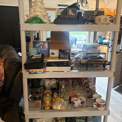Sale Photo Thumbnail #13: Four-tier plastic storage shelf holding an assortment of household goods, including a vintage white ceramic Christmas tree, glassware, mugs, electronics, and novelty items.