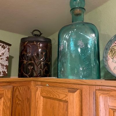 Sale Photo Thumbnail #73: Large aqua blue glass decanter bottle with cork stopper alongside a dark embossed ceramic canister set atop wooden kitchen cabinets.