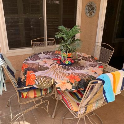 Sale Photo Thumbnail #12: Outdoor wrought iron patio dining set, featuring a round table draped with a tropical leaf tablecloth and four matching mesh-back chairs with colorful striped cushions.