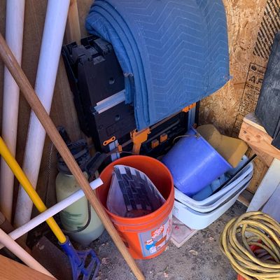 Sale Photo Thumbnail #19: Collection of assorted workshop supplies including a folded blue moving blanket, plastic buckets, long-handled tools, and a coiled extension cord in a shed corner.