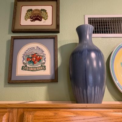 Sale Photo Thumbnail #68: Pair of framed vintage cross-stitch artwork featuring fruit label motifs alongside a tall blue glaze pottery vase.