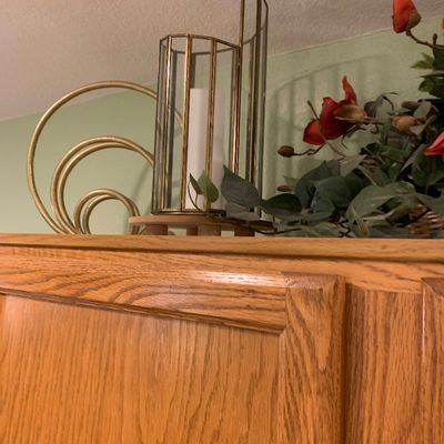 Sale Photo Thumbnail #77: Modern brass-framed glass lantern candle holder alongside decorative brass swirl art and faux floral arrangement atop an oak cabinet.