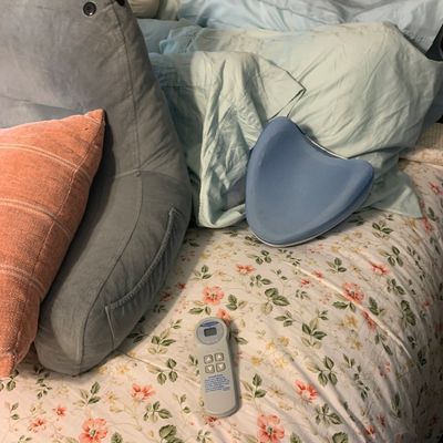 Sale Photo Thumbnail #108: A collection of bedding items including a gray backrest reading pillow, an orange throw pillow, a blue contours seat cushion, and an adjustable bed remote control resting on a floral comforter.