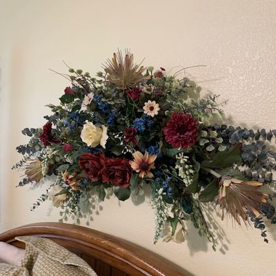 Sale Photo Thumbnail #104: Large wall-mounted artificial floral arrangement featuring deep red roses, eucalyptus, and mixed greenery, mounted above a wooden headboard.