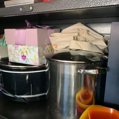 Sale Photo Thumbnail #118: Large stainless steel stockpot with glass lid sitting on a shelf alongside a black slow cooker, a floral gift box, and plastic food storage containers.
