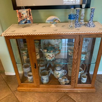 Sale Photo Thumbnail #36: Oak curio cabinet featuring glass door and side panels, housing various ceramic teapots, mugs, and pitchers, with decorative items displayed on top.