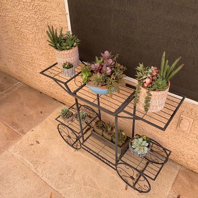 Sale Photo Thumbnail #2: Black wrought iron three-tier plant stand cart featuring decorative wheels, holding various potted artificial succulents and plants.