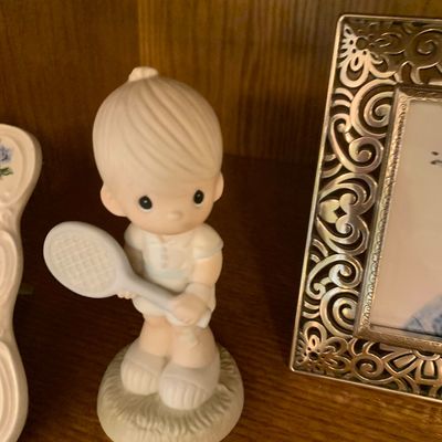 Sale Photo Thumbnail #40: Vintage Precious Moments porcelain figurine depicting a young girl holding a tennis racket, displayed on a wooden shelf next to an ornate metal picture frame.