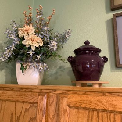 Sale Photo Thumbnail #69: Glossy plum purple ceramic jar with lid and side handles alongside an artificial floral arrangement in a square white planter.