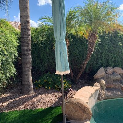 Sale Photo Thumbnail #7: Closed teal outdoor patio umbrella with fringe trim, set in a heavy round weighted base near a pool.