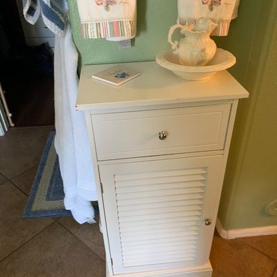 Sale Photo Thumbnail #110: White wooden bathroom storage cabinet featuring a top drawer and louvered door cabinet, topped with a decorative ceramic pitcher and basin set.