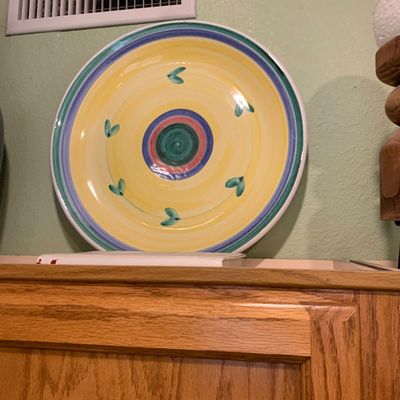 Sale Photo Thumbnail #67: Hand-painted ceramic decorative platter featuring a colorful concentric ring design in yellow, blue, teal, and pink with green leaf accents, displayed on a wooden cabinet.