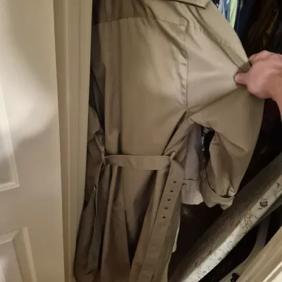 Sale Photo Thumbnail #51: Classic tan double-breasted trench coat with shoulder epaulets and a buckled waist belt, hanging in a closet.