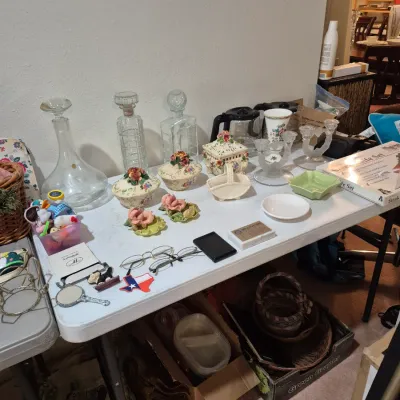 Sale Photo Thumbnail #10: A folding table displaying a variety of home goods, including glass decanters, floral porcelain containers, eyeglasses, and small decorative items.