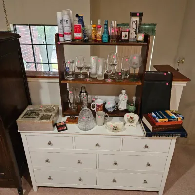 White wooden six-drawer dresser topped with a dark wood two-tier shelf displaying assorted glassware, toiletries, perfumes, ceramics, and...