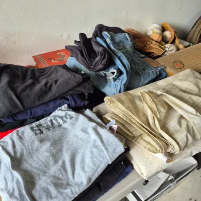 Sale Photo Thumbnail #32: Assorted folded clothing including t-shirts, denim jeans, and textiles displayed on a table alongside baseball equipment.
