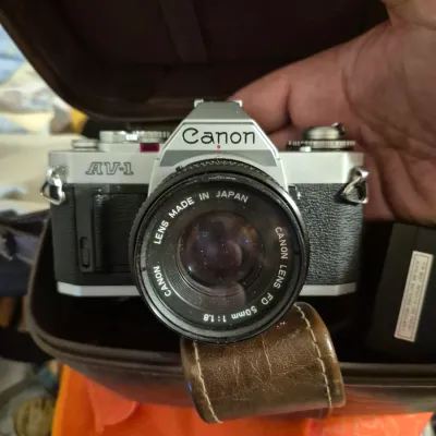 Sale Photo Thumbnail #58: Vintage Canon AV-1 35mm SLR camera fitted with a Canon FD 50mm f/1.8 lens, housed inside a fitted brown leather carrying case.