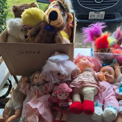 Assortment of vintage dolls, plush teddy bears, and colorful troll dolls displayed on a table and in a box.