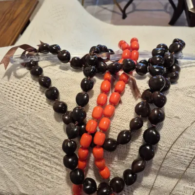 Sale Photo Thumbnail #62: Selection of beaded necklaces including polished brown kukui nut leis tied with brown ribbon accents alongside a bright orange strand.