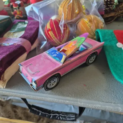 Novelty paperboard pink convertible car with surfboard, displayed alongside vintage satin Christmas ornaments and holiday decor.