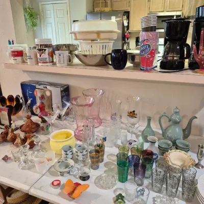 Sale Photo Thumbnail #16: Assortment of household items and glassware displayed on tables and a breakfast bar ledge, including a Black & Decker drip coffee maker, various drinking glasses, ceramic teapots, and pink glass vases.