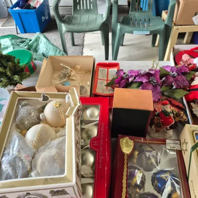 Sale Photo Thumbnail #25: Assorted vintage Christmas decor including boxed glass bauble ornaments, poinsettia flowers, and a small table display, with two green plastic patio chairs in the background.