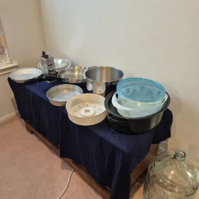Sale Photo Thumbnail #14: Collection of kitchenware including aluminum baking pans, stainless steel stockpots, a box grater, speckled enamel roaster, Pyrex bowls, and a large glass carboy jug.