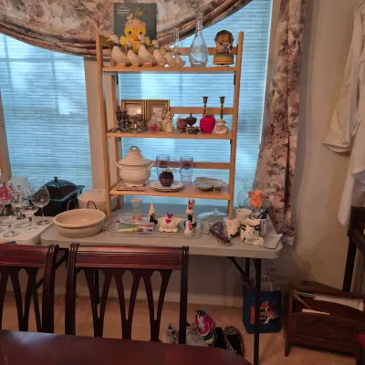 Sale Photo Thumbnail #2: A light wood four-tier shelf unit filled with home decor collectibles, including goose figurines, glassware, and brass candlesticks, arranged on a folding table.