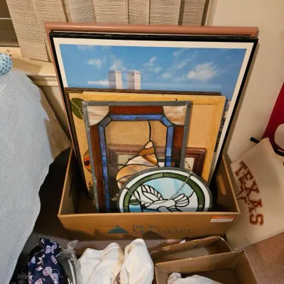 Cardboard box containing stained glass suncatchers, including a seashell and dove design, along with a framed World Trade Center photograph.