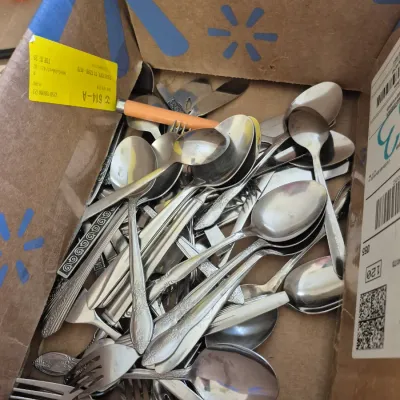 Sale Photo Thumbnail #20: Cardboard box filled with an assortment of vintage stainless steel flatware, including spoons, forks, and knives in various patterns.