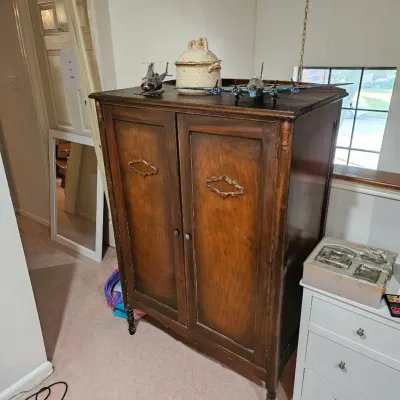 Vintage dark wood armoire featuring carved door accents, topped with decor including a model airplane, helicopter, and ceramic crock.