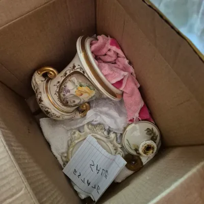 Sale Photo Thumbnail #7: Cardboard box containing vintage porcelain urns and decorative items with hand-painted floral motifs and gilt trim, wrapped in protective cloth.