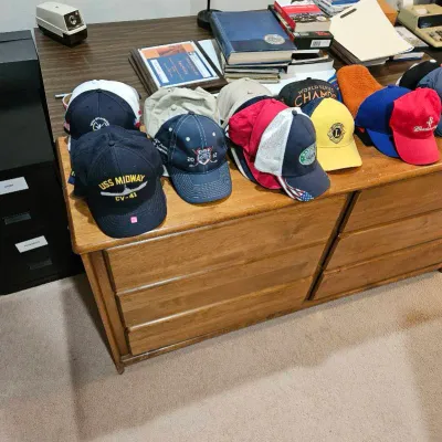 Collection of vintage baseball and trucker hats displayed on a wood dresser, including a USS Midway CV-41 cap, Lions Club hat, and...
