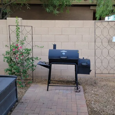 Black metal offset smoker charcoal grill, featuring a side firebox, front wooden shelf, and wheeled base for portability.