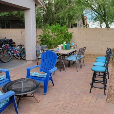 Patio furniture set including a dining table with chairs, blue plastic Adirondack style chairs, outdoor barstools, a fire pit, and bicycles.