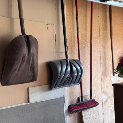 Sale Photo Thumbnail #17: Collection of long-handled yard and garage tools, including a metal scoop shovel, a plastic snow shovel, and a red push broom hanging on a garage wall in used condition.
