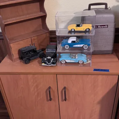 Sale Photo Thumbnail #35: Collection of die-cast model cars including Corvette convertibles in acrylic display cases alongside vintage toy cars, sat near a Bell & Howell equipment case and a wooden spice rack. Category: Toys & Hobbies / Diecast & Toy Vehicles.