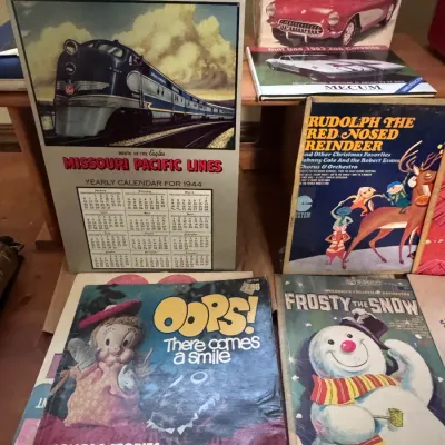 Sale Photo Thumbnail #42: Vintage memorabilia collection including a 1944 Missouri Pacific Lines train calendar, vintage Monogram '57 Corvette model kit box, Mecum auction book, and children's vinyl records like Frosty the Snowman and Rudolph the Red-Nosed Reindeer. Categories: Vi