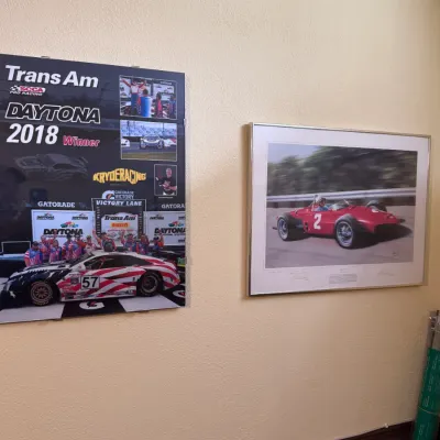 Sale Photo Thumbnail #33: Pair of framed racing wall art pieces featuring a Trans Am SCCA Pro Racing Daytona 2018 winner poster and a framed artwork print depicting a red vintage race car.