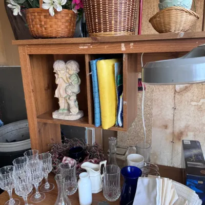 Sale Photo Thumbnail #13: Wooden desk hutch shelf displaying a children's figurine and linens, above a tabletop featuring glassware, crystal stemware, ceramics, folded cloth napkins, and woven baskets.