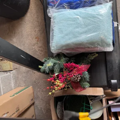 Sale Photo Thumbnail #16: Assorted household goods resting on a black exercise treadmill, featuring bagged linens and towels, a festive artificial pine floral arrangement with red ribbon and berries, and a box of cookware.