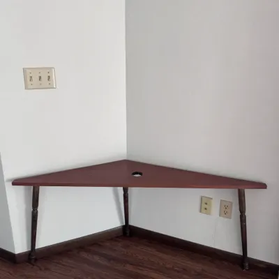 Sale Photo Thumbnail #21: Title: Triangular Wooden Corner Table with Cord Hole. Category: Furniture > Tables. Triangular reddish-brown wood corner table featuring dark turned wooden legs and a built-in circular grommet hole for cable management.