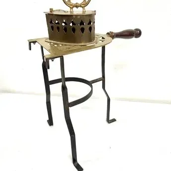Sale Photo Thumbnail #13: Antique brass coal-heated sad iron with a wooden handle, resting on a matching brass trivet supported by a tall wrought iron stand.