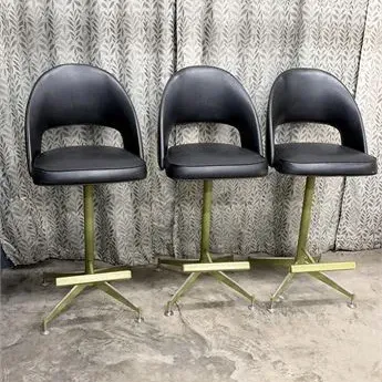 Sale Photo Thumbnail #39: Set of three mid-century modern bar stools featuring black vinyl seats with cut-out backs and brass-toned metal pedestal bases with built-in footrests.