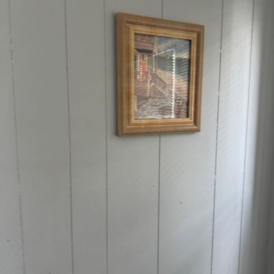 Framed original artwork depicting a coastal building and stairs, housed in a thick wood frame and displayed on a panelized wall.