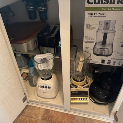 Selection of small kitchen appliances stored in a cabinet, including Osterizer blenders, a Cuisinart Prep 11 Plus food processor in its...