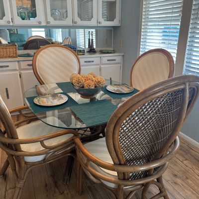 Round glass-top dining table set with four rattan armchair style chairs featuring cream upholstered seats and backs, accompanied by blue...