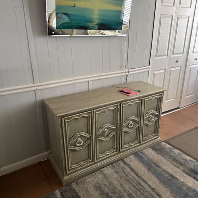 Vintage French Provincial style four-door credenza with a distressed grey finish, set beneath a wall-mounted TV featuring a mirrored bezel.